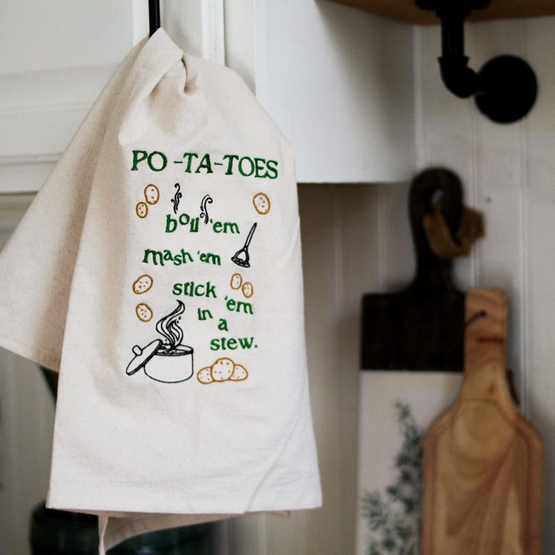 An unbleached off white linen tea towel embroidered with a quote from Lord of the Rings movie, the Two Towers. The Quote is Potatoes. Boil 'em, mash 'em, stick'em in a stew. Playful potatoes fall down to a  large whimsical pot, and a classic potatoes masher. The fonts represent the verbs, with steam coming off of the boil em phrase and the letters in mashed closely pushed together. Colors are a bold emerald green and a soft golden brown. Hang this towel in kitchen of any hobbit, elf, or wizard.