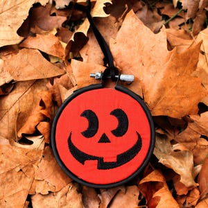 May include: A round, red Halloween decoration with a smiling jack-o'-lantern face embroidered in black thread. The embroidery hoop is black, with a silver metal clasp and a black ribbon for hanging. The decoration rests on a bed of dried, brown leaves.