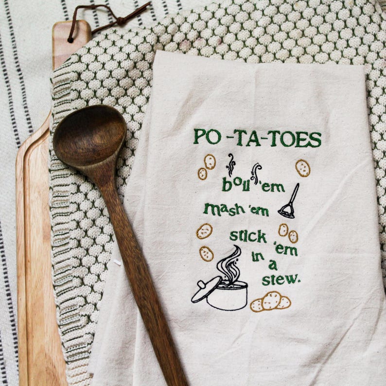 An unbleached off white linen tea towel embroidered with a quote from Lord of the Rings movie, the Two Towers. The Quote is Potatoes. Boil 'em, mash 'em, stick'em in a stew. Playful potatoes fall down to a  large whimsical pot, and a classic potatoes masher. The fonts represent the verbs, with steam coming off of the boil em phrase and the letters in mashed closely pushed together. Colors are a bold emerald green and a soft golden brown. Hang this towel in kitchen of any hobbit, elf, or wizard.