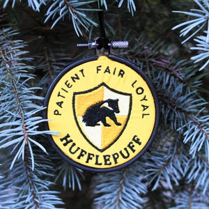 May include: A yellow and black embroidered ornament with the Hufflepuff house crest from the Harry Potter series. The crest features a badger and the words "Patient, Fair, Loyal, Hufflepuff".
