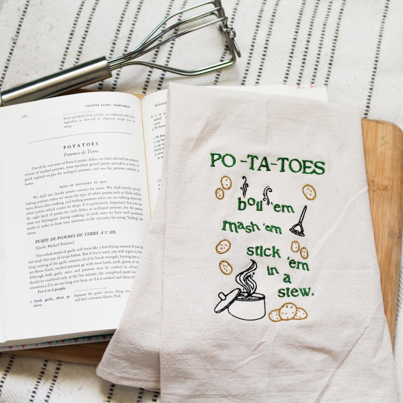 An unbleached off white linen tea towel embroidered with a quote from Lord of the Rings movie, the Two Towers. The Quote is Potatoes. Boil 'em, mash 'em, stick'em in a stew. Playful potatoes fall down to a  large whimsical pot, and a classic potatoes masher. The fonts represent the verbs, with steam coming off of the boil em phrase and the letters in mashed closely pushed together. Colors are a bold emerald green and a soft golden brown. Hang this towel in kitchen of any hobbit, elf, or wizard.