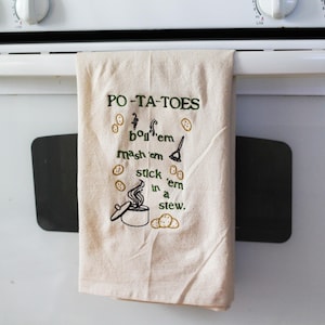 An unbleached off white linen tea towel embroidered with a quote from Lord of the Rings movie, the Two Towers. The Quote is Potatoes. Boil 'em, mash 'em, stick'em in a stew. Playful potatoes fall down to a  large whimsical pot, and a classic potatoes masher. The fonts represent the verbs, with steam coming off of the boil em phrase and the letters in mashed closely pushed together. Colors are a bold emerald green and a soft golden brown. Hang this towel in kitchen of any hobbit, elf, or wizard.