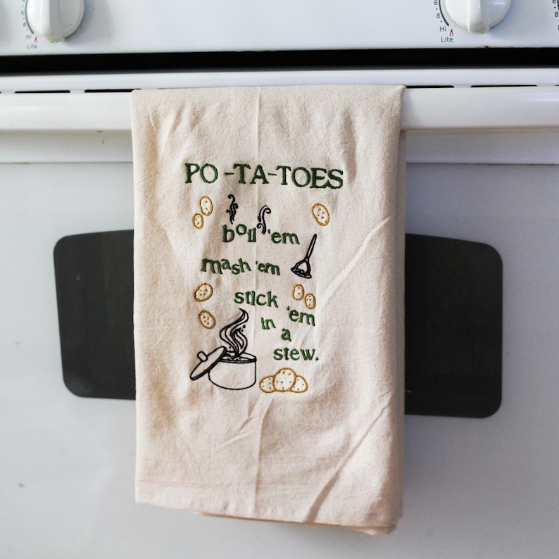 An unbleached off white linen tea towel embroidered with a quote from Lord of the Rings movie, the Two Towers. The Quote is Potatoes. Boil 'em, mash 'em, stick'em in a stew. Playful potatoes fall down to a  large whimsical pot, and a classic potatoes masher. The fonts represent the verbs, with steam coming off of the boil em phrase and the letters in mashed closely pushed together. Colors are a bold emerald green and a soft golden brown. Hang this towel in kitchen of any hobbit, elf, or wizard.