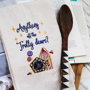 May include: A cream-coloured tea towel embroidered with the words "Anything off the Trolly, dears?" in blue script, with gold stars and colourful candy and luggage designs. A wooden spoon with a geometric design rests beside it.