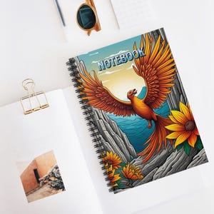 May include: Spiral-bound notebook featuring a vibrant illustration of a phoenix with outstretched wings, set against a backdrop of a mountain range, ocean, and sunflowers. The word "NOTEBOOK" is printed across the top. The notebook is open on a white surface.