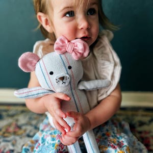May include: A handmade stuffed bear doll with a pink bow and ears. The bear is white with blue and pink stripes. The doll has a stitched brown nose and black eyes. The child is wearing a floral skirt and a light-colored top.