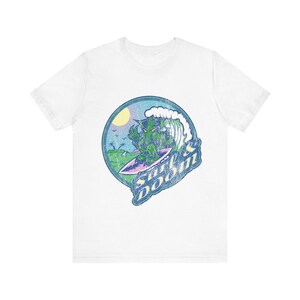 May include: White t-shirt featuring a circular graphic of a green creature surfing a wave. The design includes a sun, palm trees, and the text "Surf & Doom" in a distressed font. The shirt is made of a soft material.