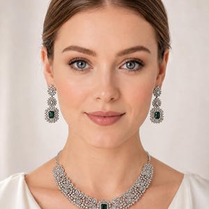 Op de afbeelding: Een zilverkleurige sieradenset bestaande uit een ketting en bijpassende oorbellen. Elk sieraad heeft een centrale smaragdgroene rechthoekige steen, omringd door een halo van kleine heldere steentjes en gedetailleerd zilverkleurig filigraan.