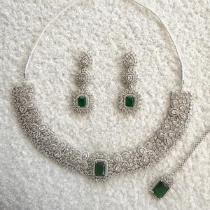 May include: A silver-toned jewelry set featuring a necklace, earrings, and a pendant. The necklace and earrings have a floral design with clear stones and a central emerald-green rectangular stone. The pendant mirrors the design of the earrings.
