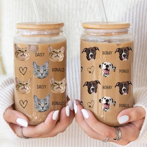 Op de afbeelding: Twee glazen bekers met houten deksels en doorzichtige rietjes. Eén heeft kattengezichten, namen en harten; de andere heeft hondengezichten, namen en harten. Beide zijn gevuld met een lichtbruine vloeistof.