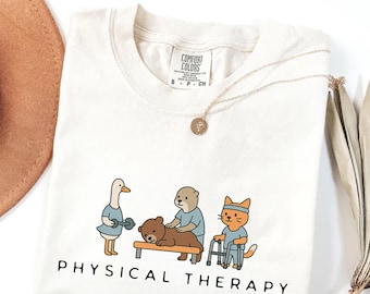 May include: Cream-colored t-shirt with a "Physical Therapy" graphic. The graphic features cartoon animals: a duck lifting weights, a bear receiving a massage, and a cat using a walker. A gold necklace with a pendant is draped on the shirt.
