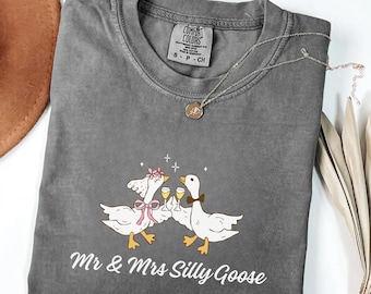 May include: A gray Comfort Colors t-shirt with a whimsical graphic of two geese toasting with champagne glasses. The text "Mr & Mrs Silly Goose" is printed below the illustration. A silver necklace with a pendant is draped on the shirt.