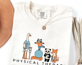May include: Cream-colored t-shirt with a whimsical illustration of animals engaged in physical therapy. A goose with crutches, a raccoon on a balance ball, a panda with a clipboard, and a fox. The words "PHYSICAL THERAPY" are printed below the illustration.