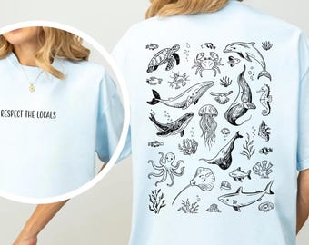 May include: Light blue t-shirt with a black and white illustration of various sea creatures on the back, including a turtle, whale, and shark. The front of the shirt has the text "Respect the Locals" in black.