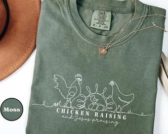 May include: A moss green t-shirt with a white line drawing of a rooster, hen, and chick, with the text "CHICKEN RAISING and Jesus praising". A silver necklace with a small pendant rests on the shirt.
