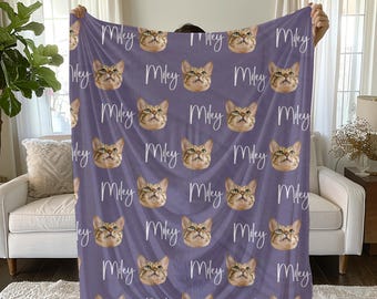 May include: A purple blanket featuring a repeating pattern of a cat's face and the name "Miley" in white script. The cat has brown and tan fur. The blanket is held up in front of a white couch and a window.