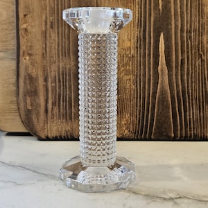 May include: A clear, faceted glass candlestick holder with a textured, cylindrical body and a hexagonal base. The top is a wider, flat surface. The background includes a wooden cutting board.