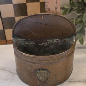 May include: A semi-circular wall-mounted copper container with a weathered patina and a lion head ring pull on the front. A wooden ruler placed behind the item indicates a height of approximately 6 inches from the base to the top edge.