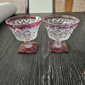 May include: Two clear glass dessert dishes with a pink rim and a diamond pattern. The dishes have a square base and are sitting on a black surface.