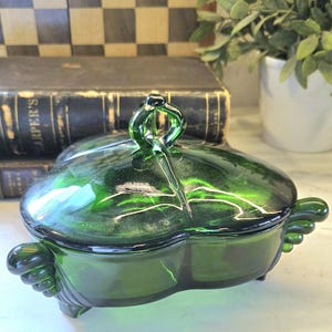 May include: A green glass trinket dish with a lid. The dish is heart-shaped with a loop handle on the lid and decorative feet. The glass has a glossy finish. The background includes books and a plant.