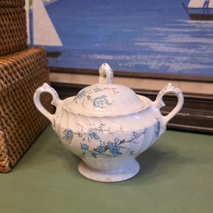 May include: White ceramic sugar bowl with a lid, featuring blue floral patterns. The bowl has two handles and a pedestal base. The background includes a painting of a seascape and a woven basket.