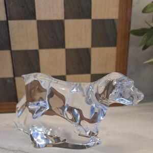 May include: A clear glass dog figurine, sculpted in a seated position. The figurine is highly reflective, with light refracting through the glass. The background includes a checkered pattern and a wooden frame.