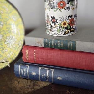 May include: A vintage ceramic cup with a floral and architectural design, resting on a stack of books. The books have red, gray, and blue covers. A yellow plate with a floral pattern is visible on the left.