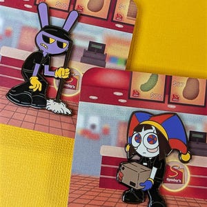 Op de afbeelding: Twee emaille pins met cartoonfiguren in een winkelomgeving. Eén pin toont een paars konijn met een bezem, gekleed in een zwarte outfit en gele handschoenen. De andere pin toont een personage met een narrenhoed die een doos vasthoudt. Beide pins hebben een glanzende afwerking.