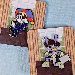 Peut inclure: Deux épingles en émail représentant des personnages de dessins animés. L'une représente une personne en costume bleu et chapeau de bouffon, tenant une tablette. L'autre montre un lapin violet portant un chapeau de cow-boy et tenant une mallette. Les deux épingles sont sur un fond de fenêtre et de rideaux.