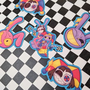 Op de afbeelding: Een verzameling kleurrijke cartoonstickers met nar-achtige personages met harlekijnhoeden en konijnenoren. De stickers hebben een blauwe rand en staan op een zwart-wit geblokte achtergrond.