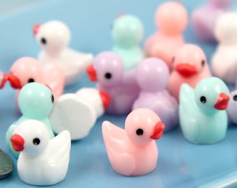 May include: A collection of miniature duck figurines in various pastel colors, including pink, purple, and blue, with red beaks and black eyes. The ducks are arranged on a light blue rectangular tray, with a coin visible in the foreground.