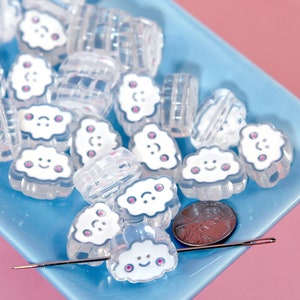 Könnte beinhalten: Klare, wolkenförmige Perlen mit lächelnden Gesichtern und rosa Akzenten, in einer hellblauen Schale. Eine Nadel und ein Penny liegen ebenfalls auf einem rosa Hintergrund.