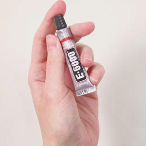 May include: A small silver metal tube of E-6000 adhesive featuring a black screw-on cap, a red horizontal stripe, and a black label with white text reading E-6000. The tube has a crimped bottom edge and is held upright against a plain background.