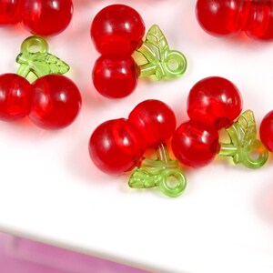May include: A collection of red plastic cherry charms with green plastic stems and leaves. The charms are approximately the size of a UK ten pence coin.