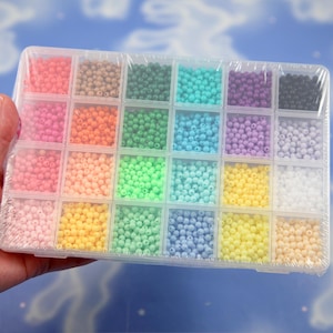 May include: A clear plastic rectangular storage container with twenty-four individual compartments, each filled with small round beads in a variety of solid colors including pink, orange, green, blue, purple, yellow, white, and black.