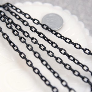 May include: Five strands of black chain, likely for jewelry making, are laid out on a white surface. The chains are made of small, oval-shaped links.