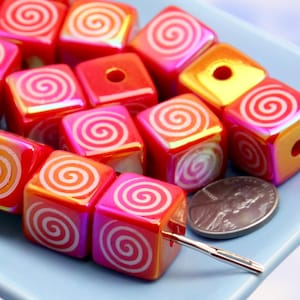 May include: Close-up of square beads in shades of red, pink, and orange, each with a white spiral design. The beads are strung on a needle, with a penny for size comparison. The beads have a glossy finish.