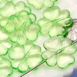 May include: Close-up of translucent green clover-shaped beads. The beads are strung on a silver needle, and a coin is visible for size comparison. These beads are suitable for jewellery making and craft projects.