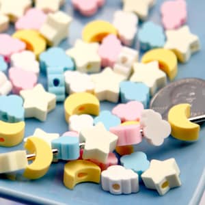 May include: A selection of pastel-coloured beads in star, moon, and cloud shapes. The beads are in yellow, pink, blue, and white. Some beads are strung on a metal rod, and a coin is visible in the background.