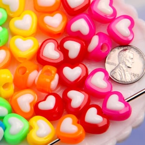 May include: A collection of colorful heart-shaped beads in various colors, including blue, green, yellow, orange, red, and pink, each with a white heart design. A US penny is included for size reference. These beads are likely for jewelry making or crafts.