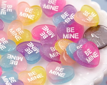 May include: A collection of colorful heart-shaped candies with the words "Be Mine" printed on them. The candies are translucent and have a glittery finish.