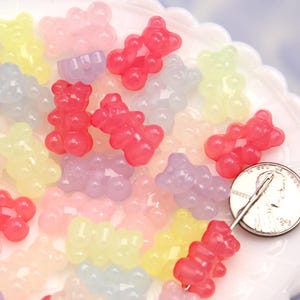 Könnte beinhalten: Eine Sammlung von bunten, gummibärchenförmigen Perlen in verschiedenen transparenten Farben wie Rot, Rosa, Gelb, Lila und Blau. Die Perlen sind auf einer weißen Oberfläche verstreut, mit einer Münze als Größenvergleich. Ideal zum Basteln.
