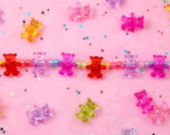 May include: A string of colorful gummy bear charms on a pink background with iridescent glitter. The gummy bears are in various colors, including red, orange, yellow, green, blue, purple, and pink.