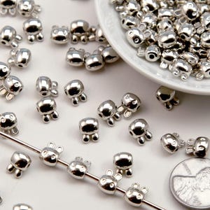 May include: A collection of small, silver-colored metal beads shaped like rabbit heads. The beads are scattered on a white surface, with many contained in a small white bowl. A metal needle is threaded with five beads, and a US dime is shown for scale.