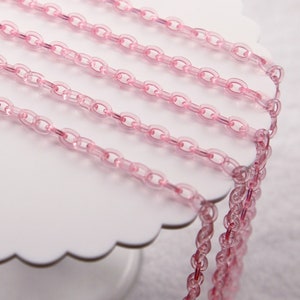 May include: Pink plastic chain links arranged on a white scalloped-edge cake stand. A U.S. quarter is next to the chain. The chains are made of translucent pink plastic. The chains are arranged in parallel lines.