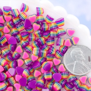 以下が含まれることがあります： カラフルなハート型の樹脂ビーズの山をクローズアップした写真です。ビーズはピンクとパープルのミックスで、レインボーストライプが施されています。スケールのためにアメリカ1セント硬貨が添えられています。