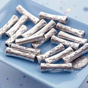 May include: Silver-colored metal tube beads with etched spiral and cross designs. The beads are cylindrical with a hole through the center. They are displayed on a light blue square plate.