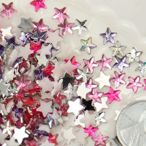 May include: A close-up of a variety of star-shaped rhinestones in silver, pink, and purple. The rhinestones are scattered on a white surface and a US quarter is included for scale.
