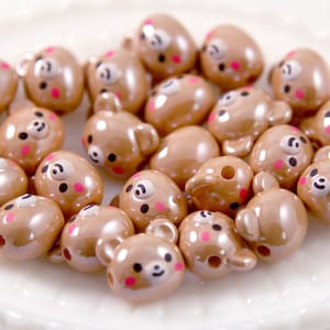 May include: A collection of brown bear-shaped beads with black eyes, small ears, and pink cheeks. The beads have a glossy finish and a hole for stringing. These decorative beads are ideal for jewelry making and crafting projects.