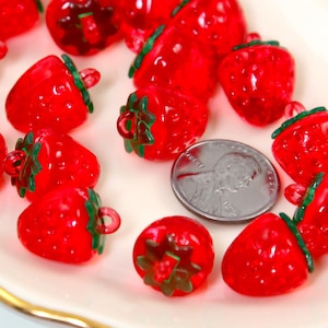 May include: A collection of translucent red strawberry-shaped beads with green stems and a silver coin. The beads are scattered on a white and gold-rimmed plate. The beads are approximately 0.75 inches in length.
